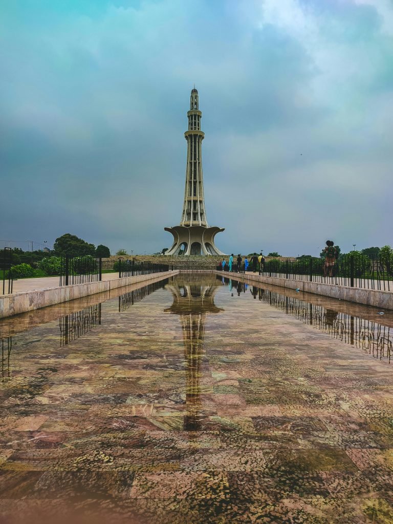 Stunning vertical capture of Minar-e-Pakistan in Lahore with its reflection in water, under a cloudy sky.